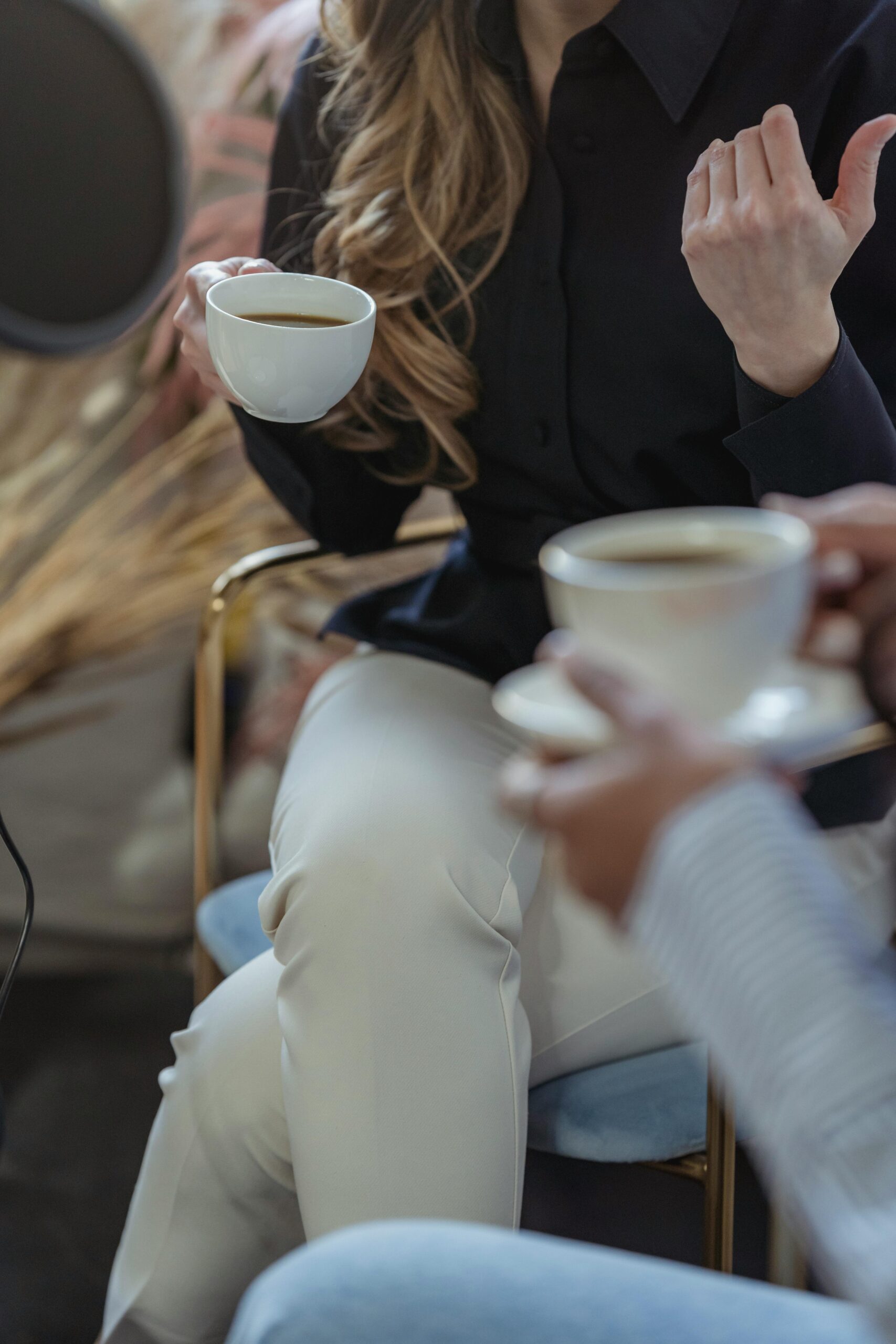 Two people are sitting and holding white coffee cups. The person on the left is wearing a black shirt and white pants, gesturing with their hand. The background is softly blurred, and theres a microphone partially visible on the side.