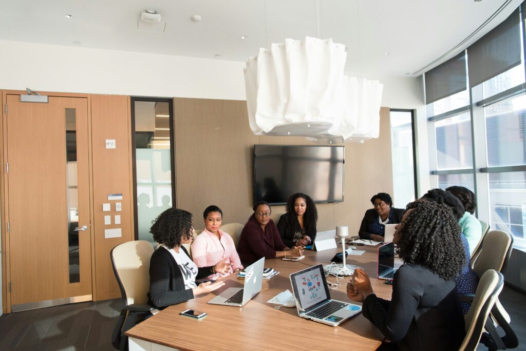 A group of women in leadership sit around a conference table in a modern office. Laptops are open as they engage in discussion. A large screen is on the wall, and a contemporary light fixture hangs above. Large windows let in natural light. One of the topics on their agenda, the mental health of their employees.