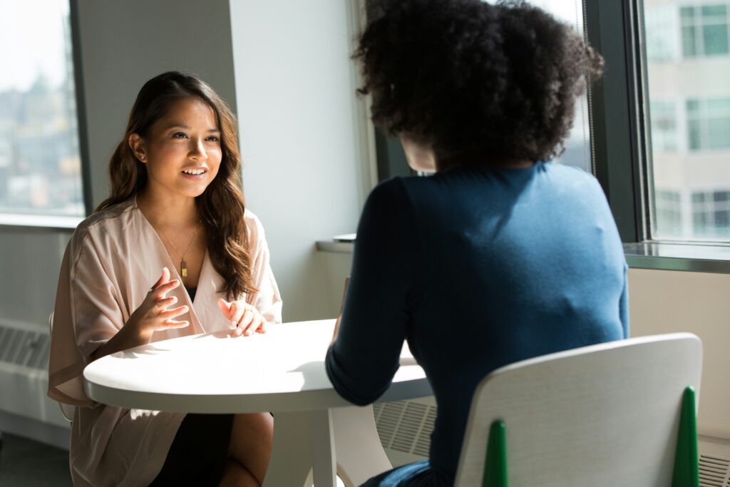 Two women are sitting at a small round table in a bright room. One woman is speaking and gesturing with her hands, while the other listens attentively. They are near a window with city buildings visible outside.