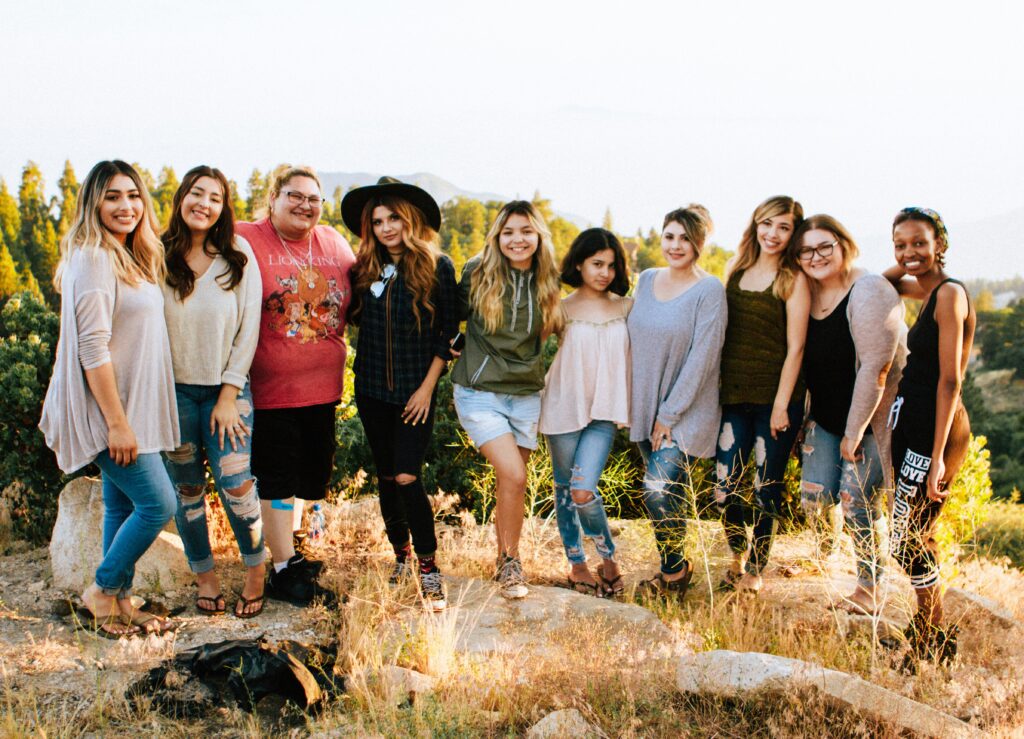 A Team of women on a retreat stand on a trail in the wilderness. They are diverse in culture, background, age, shape, size, and skin tone. Their Emerging Leader fosters an inclusive and inspiring culture.