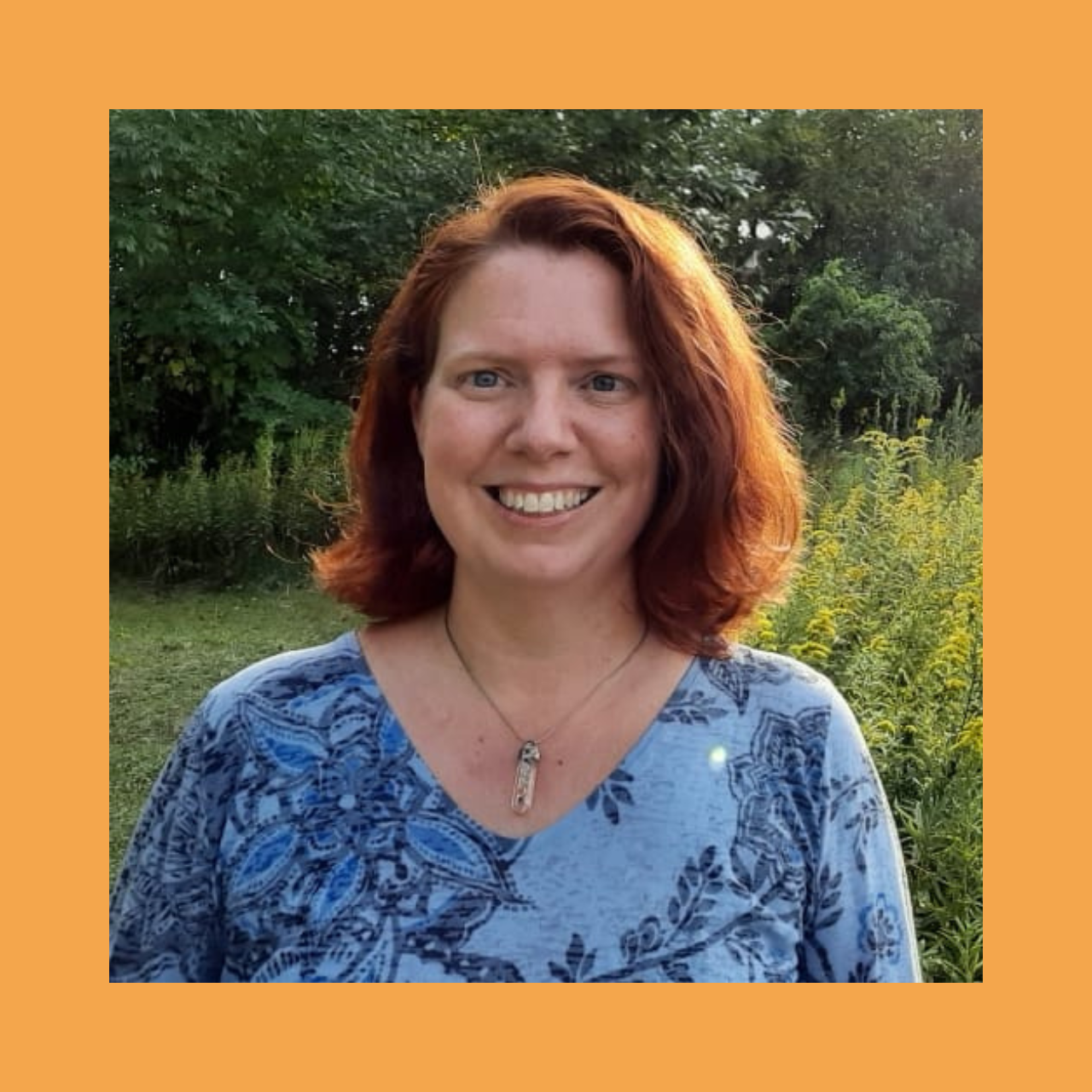 A person with shoulder-length red hair smiling, wearing a blue patterned top and a necklace with a pendant. Behind them is a garden with yellow flowers and green trees.