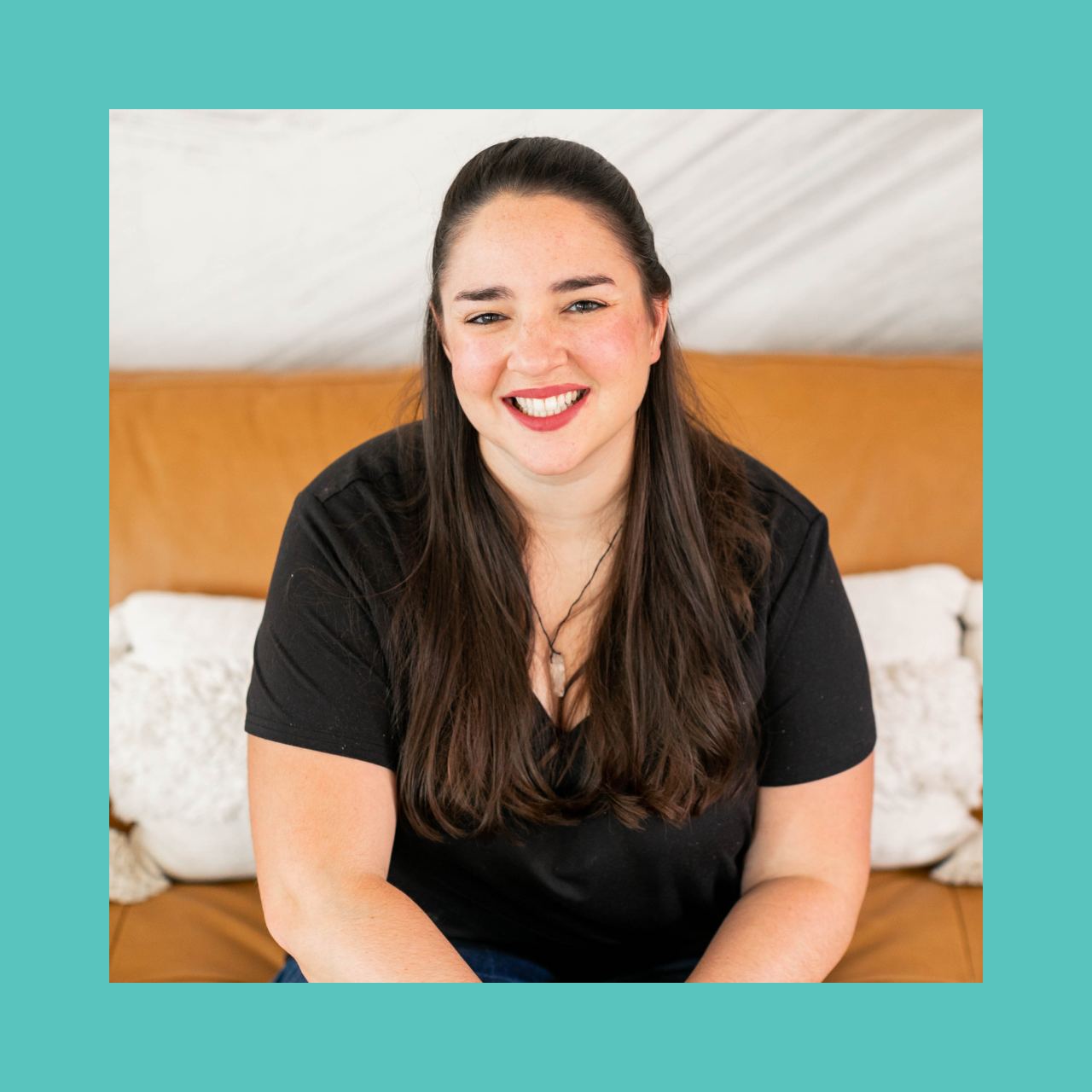 A smiling person wearing a blue polka dot scarf stands outdoors. They have sunglasses atop their head, and a patterned shirt. The background features a building with large windows.
