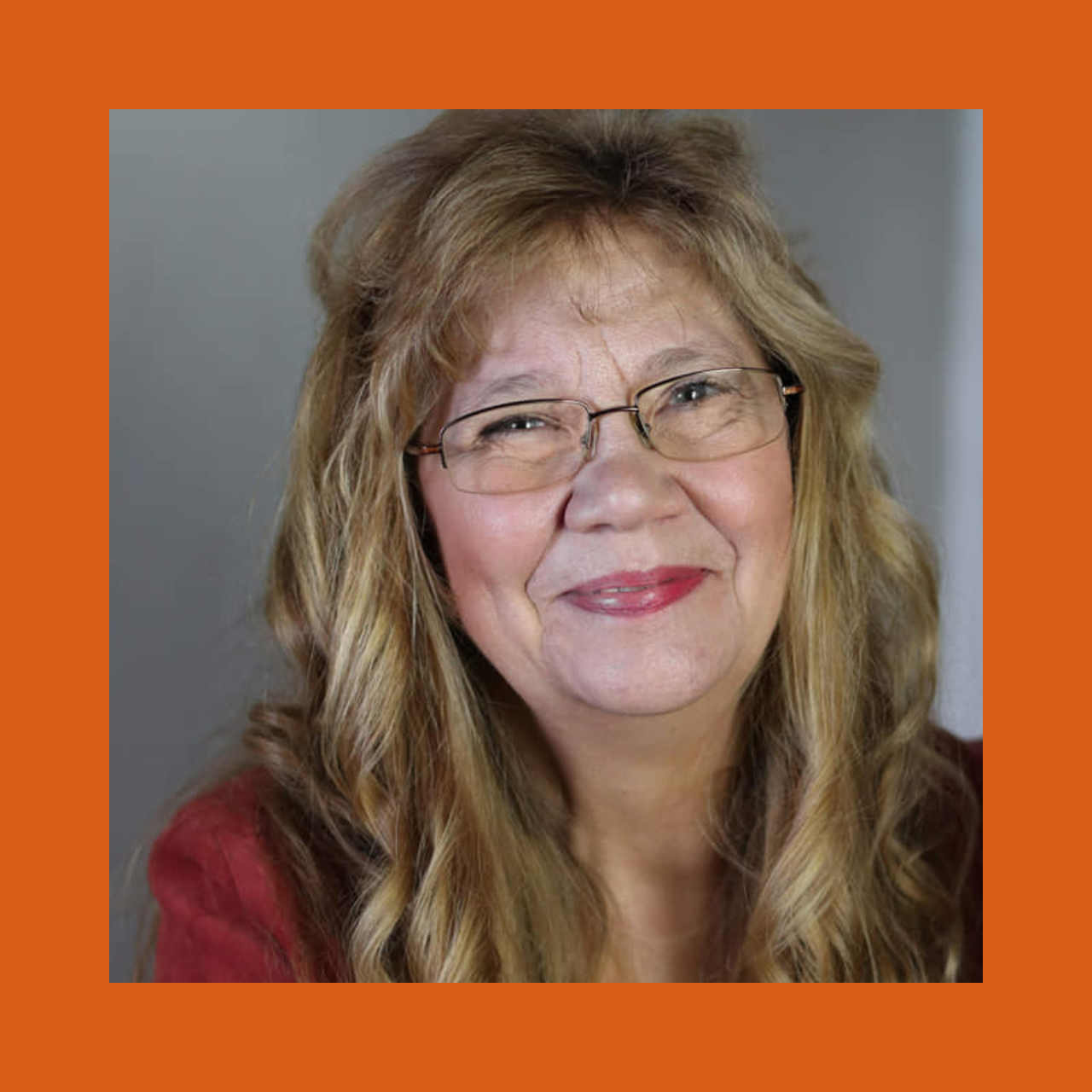 Smiling woman with wavy, long blonde hair and glasses, wearing a red top, set against a gray background framed by a pink border.